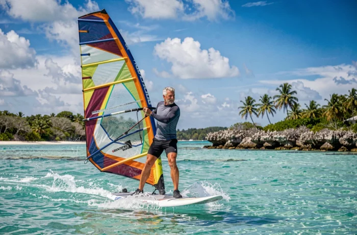 Légende du windsurf français sur son board dans un lagon tropical, voile colorée au vent, eau turquoise et récif corallien en arrière-plan